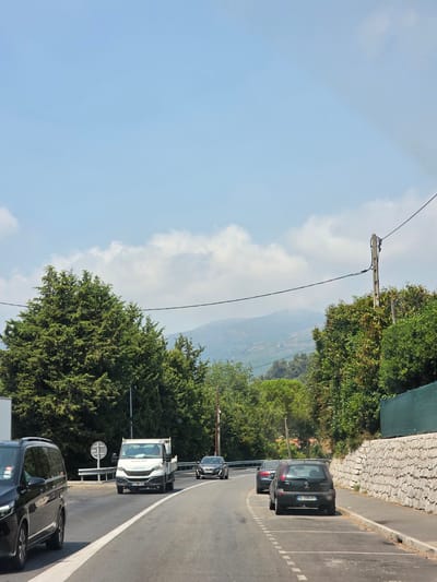 This is a photograph taken from a vehicle traveling on a road in Vence, France. The road curves to the right and is bordered by trees on both sides and a stone wall on the right. Several cars are visible on the road, including a black minivan in the foreground on the left, a white Iveco delivery truck in the middle lane, and a dark sedan behind it. In the distance, hills covered in trees and vegetation are visible under a bright blue sky with scattered clouds. The lighting suggests it is daytime, likely midday, given the brightness and the shadows.