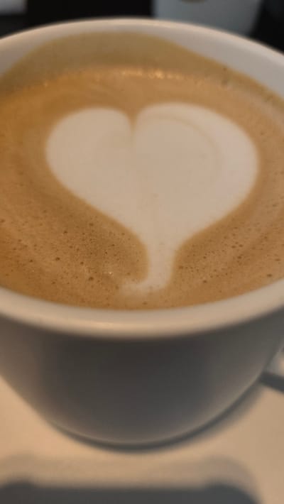 The image shows a close-up of a white mug filled with coffee, featuring latte art in the shape of a heart. The coffee has a foamy crema with small bubbles, indicating it is likely a latte or cappuccino. The mug is positioned on a light-colored surface, possibly a saucer or table. The lighting suggests an indoor setting, possibly a cafe. The image does not provide any specific visual cues that would allow for the identification of a location within Gwangmyeong, South Korea.