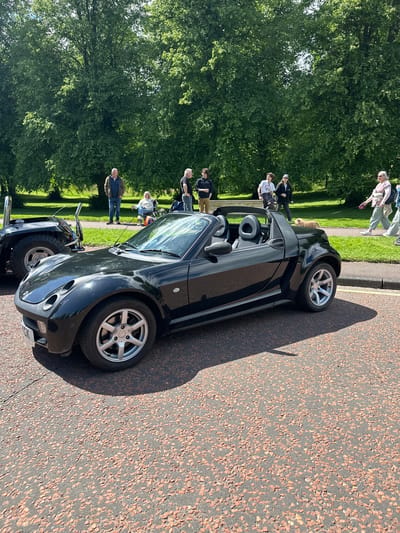 A black convertible smart car is parked on a tree-lined street in Belfast City, United Kingdom. The car is positioned facing right, with its headlights visible. In the background, several people are standing and walking, some near large trees. The street is paved with asphalt containing red chippings. The scene appears to be outdoors on a sunny day.