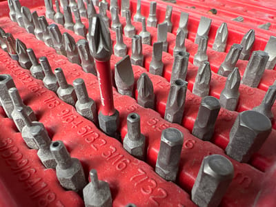 The image shows a close-up, high-angle view of a red tool organizer filled with various screwdriver bits. The bits are mostly metallic gray and are arranged in rows within the red plastic. Many of the bits have hexagonal bases, and some have visible markings indicating their size and type, such as "3/16", "5/32", and "7/32". One prominent bit, to the left of the center, is red and appears to be a Torx bit. The organizer itself has a textured surface and some dust is visible on the bits and the organizer, suggesting use. The background is out of focus but appears to be the continuation of the tool organizer. There are no people or animals present in the image. The scene does not contain any elements that would allow for the localization of the city of Lanžhot, Czechia.