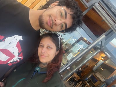 A daytime selfie photograph depicts two individuals, a male and a female, positioned in the foreground. The background reveals the exterior and interior of a commercial building in Tolhuin, Argentina.
The male, situated to the left, has dark, curly hair, a beard, a mustache, and wears gold-framed eyeglasses. He exhibits a septum piercing and multiple ear piercings. He is dressed in a black t-shirt featuring a white Ghostbusters logo. His expression is a slight smile.
The female, positioned to the right, has reddish-brown hair and visible facial piercings, including a septum ring and a nostril stud. Intricate black ink tattoos are visible on her neck, extending to her collarbone, and partially on her right forearm. She wears a dark green fleece jacket. Her expression is also a slight smile.
The background consists of the building's facade, featuring light-colored wooden slats at the top. Below, large glass panels framed in dark material provide a view into the interior. Reflections on the glass show elements of the sky and internal lighting. Inside, wooden walls and ceiling are visible, along with multiple dark chairs, wooden tables, and pendant light fixtures. A prominent banner or poster with Argentine flag colors and the text "ARA SAN JUAN" is displayed inside the establishment. Other smaller Argentine flag elements and logos, including a light bulb graphic, are also visible through the glass. The scene is located in Tolhuin, Argentina, indicating a public or commercial setting such as a cafe or restaurant.