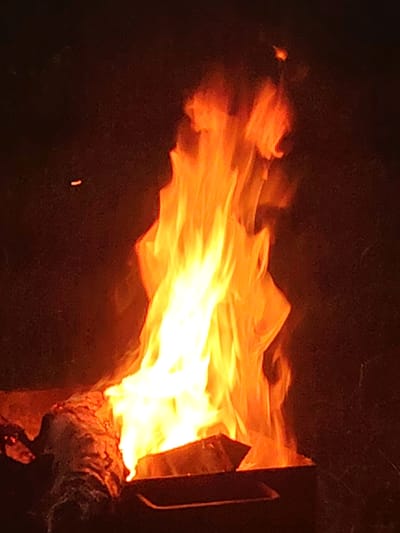 The media file shows a close-up shot of a campfire at night. The flames are a vibrant mix of orange, yellow, and white, creating a bright and dynamic display against the dark background. In the foreground, a log is partially visible, charred and contributing to the fire. The fire appears to be contained within some sort of metal fire pit or grill. The overall scene evokes a sense of warmth and the primal comfort associated with a bonfire. The lighting is dominated by the fire, casting an orange glow and creating deep shadows. The time of day is clearly night due to the darkness of the surroundings. There is no visible text in the media file. The location context, Заямнаўскі сельскі Савет, Belarus, suggests an outdoor setting, possibly in a rural area or park. The image does not contain any people, and the focus is solely on the natural phenomenon of the burning fire and the wood.