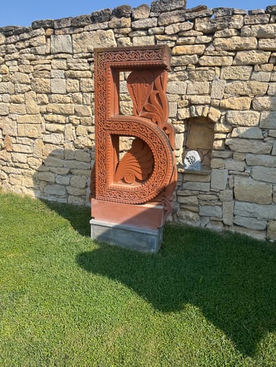 This image showcases an ornately carved stone monument, likely a khachkar given the location context of Pliska, Bulgaria. The monument is crafted from reddish-brown stone and features intricate geometric patterns and stylized bird motifs. It is positioned in front of a rough-hewn stone wall, creating a juxtaposition of architectural styles. To the right of the monument, a vibrant green lawn stretches out, casting a distinct shadow, indicating the presence of sunlight and suggesting it is a sunny day. The stone wall appears to be part of an ancient structure, possibly ruins, with some sections showing signs of age and weathering. A small, circular decorative element is visible embedded in a niche within the stone wall. The overall scene suggests an outdoor setting, likely a historical or archaeological site. The image is captured from a slightly elevated perspective, looking downwards. There is no visible text in the image.