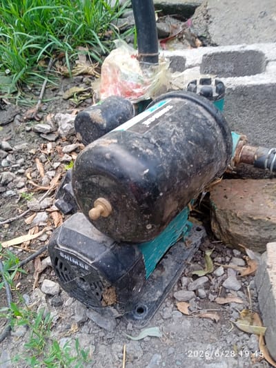 A close-up, high-angle shot captures a water pump with a black motor and a black tank situated on the ground. The pump is partially obscured by debris, including rocks, dirt, and dried leaves. Green grass is visible in the upper left corner, and a concrete block structure is present in the background. A watermark in the lower right corner indicates the date and time as 2026/6/28 19:45. The scene appears to be outdoors, possibly in a yard or an area with construction or disused equipment.