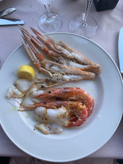 A white plate of seafood rests on a light pink tablecloth. The plate contains langoustines, shrimp, and what appears to be calamari. A wedge of lemon is also on the plate, along with a small amount of oil and black pepper. Two wine glasses are visible in the background, and cutlery is present on either side of the plate. The scene appears to be an outdoor dining setting, possibly a restaurant in Pozzarello, Italy, given the presence of seafood and wine glasses. The lighting suggests it is daytime.