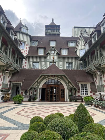 The exterior courtyard of the Hôtel Le Normandy in Deauville, France is depicted. The hotel exhibits traditional Normandy architecture with white half-timbering and green trim. The courtyard features a patterned paved ground with a central octagonal design of red lines on a beige background, bordered by grey pebble pathways. In the foreground, meticulously manicured boxwood bushes are arranged in various rounded and conical shapes. To the right, a row of bicycles with green frames is parked along the building. Benches and potted plants are placed near the entrance of the hotel, which features large wooden doors under an arched walkway with the hotel's name, "Le Normandy," inscribed above. The sky is overcast.