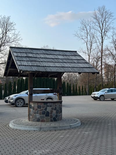 This image displays a paved parking area in Koknese, Latvia, under a clear, light blue sky with a few wispy white clouds. A central, decorative well structure occupies the foreground, situated on a circular, raised island of light grey paver blocks. The well features a dark grey wooden shingled roof supported by four substantial wooden posts. Its cylindrical base is constructed from an array of multi-colored fieldstones, and a wooden crank mechanism is visible on its right side.
The surrounding ground is uniformly covered in light grey paver blocks laid in a herringbone pattern. In the mid-ground, two light-colored sport utility vehicles are parked. A silver SUV, identifiable as a Nissan Qashqai or similar model, is positioned to the left of the well, facing left. To the right, a white Dacia Duster SUV is parked, oriented to the right.
The background features a linear hedge composed of slender, dark green coniferous trees. Beyond this hedge, numerous bare deciduous trees with visible branches indicate a late autumn or early spring season. No individuals are visible within the scene. The lighting suggests daytime conditions.