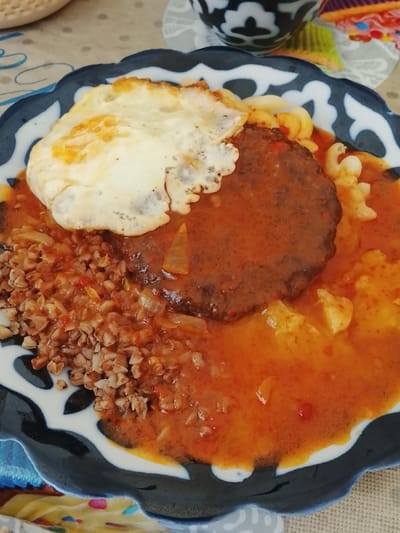 Here's a detailed description of the image:
**Scene:** A plate of food is presented on a blue and white patterned plate.
**Subjects & Objects:**
* **Food:** The meal consists of a meat patty covered in a reddish-brown sauce. A fried egg sits atop the patty. The plate also contains portions of cooked buckwheat with sauce, and some macaroni.
* **Plate:** The plate features a distinctive blue and white design.
* **Background:** A table surface is visible in the background along with a cup.
**Environment:**
* The setting appears to be indoors, potentially in a home or restaurant.
**Location Clues:** There is an item that is typically manufactured in Uzbekistan. It is an ornately decorated bowl that may be used for serving. The bowl in the image is of a similar design to the bowls that come from Uzbekistan, and that is the likely location of the image. Without more information, it is impossible to determine what the specific location is in the city of Syrdarya, Uzbekistan.