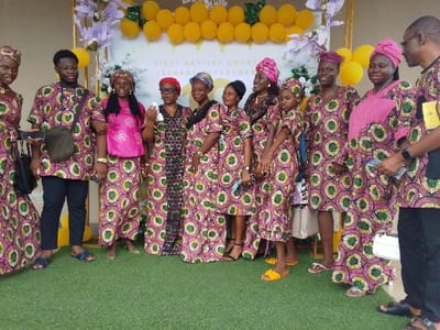 The image shows a group of nine people standing in front of a building in Yenagoa, Nigeria. The building has a sign that says "First Baptist Church Teenager Department". The people are mostly women, all dressed in colorful traditional clothing with a pink base and a green, black, and yellow pattern. The men are wearing similarly patterned shirts and trousers. One man is wearing a black shirt and trousers. They are standing on green turf, and there are yellow and purple balloons in the background. The people are all looking at the camera, some smiling and some with neutral expressions. Some of them are holding purses or bags. The scene appears to be a gathering of people in celebration of a church event.