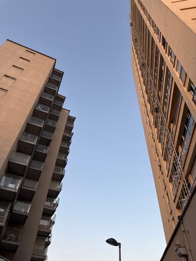 This is a low-angle shot of two modern apartment buildings, viewed from below against a clear blue sky. The perspective is disorienting, with the buildings appearing to lean in towards each other. The building on the left is much taller and appears to be a hotel or a large residential block with numerous floors featuring balconies. The building on the right is shorter and has a terraced design with repeating balcony structures.

A street lamp is visible in the upper right quadrant of the image, attached to a pole extending from a dark horizontal structure that could be a roof or another building. The lighting suggests it is daytime, likely late afternoon or early morning due to the warm tones of the buildings and the clear sky. There is no visible text within the image. The scene appears to be a contemporary urban or resort environment in Platja D'aro, Spain, characterized by modern architecture. The overall mood is serene, with the clear sky and clean lines of the buildings creating a sense of order and calm.