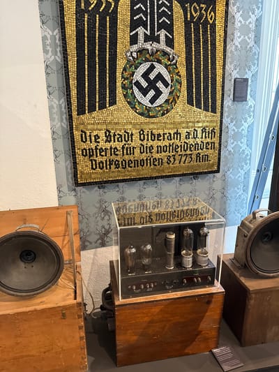 The image shows a mosaic banner with a swastika in the center, framed in black and gold. Above the swastika is the year 1936. To the right of the swastika, the banner says in gold lettering, "Die Stadt Biberach a.d. Riß opferte für die notleidenden Volksgenossen 83 773 km." To the right of the banner are two vintage loudspeakers in wooden boxes, one above the other. In between these two loudspeakers is a brown wooden box containing vacuum tubes, protected by a clear plastic case. Next to the top loudspeaker is a plaque containing information about the objects. The background is a patterned wallpaper.