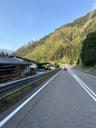 The image shows a paved road with double white lines in the foreground, indicating a two-lane highway. The road is flanked by a metal guardrail on the left and a stone-lined embankment on the right. On the left side of the road, there is a building with a dark, sloped roof, and a stack of lumber is visible next to it. Further down the road, two cars are driving, and the road curves to the right, leading into a mountainous area covered in pine trees. The sky is clear and blue. Based on the context provided by other images in the set, this scene is located in Predazzo, Italy.
