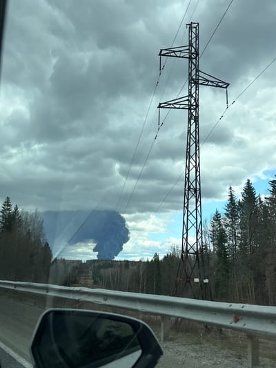 The image, taken from a moving vehicle in what appears to be Petushki, Russia, captures a roadside scene with a prominent electricity pylon and a large, dark plume of smoke in the distance.
In the foreground, on the right side, a grey metal guardrail runs horizontally along the edge of the road, partially obscuring the view of the ground. Below the guardrail, a black car side mirror is visible in the bottom right corner.
Dominating the left side and extending across the midground is a tall, dark-colored lattice-steel electricity pylon, supporting multiple power lines that stretch across the frame. The pylon's intricate metal structure is clearly visible against the sky.
The sky above is largely overcast with heavy, grey clouds, creating a subdued light. However, toward the right and further into the distance, patches of brighter blue sky are visible between the clouds.
Beyond the pylon and guardrail, a dense forest lines the horizon. Many trees, especially closer to the foreground, appear to be deciduous and bare, suggesting a late winter or early spring season. Coniferous trees are also present.
In the far distance, against the clearer blue sky, a massive, dark grey to black cloud or plume of smoke billows upwards, suggesting a significant fire or industrial event. This plume is the most striking feature in the background, contrasting sharply with the pale sky and clouds.
While two light-colored rectangular shapes, possibly signs, are visible attached to a distant power structure behind the trees, no text is legible due to the distance and image resolution. The scene depicts a daytime setting, likely on a highway or rural road.