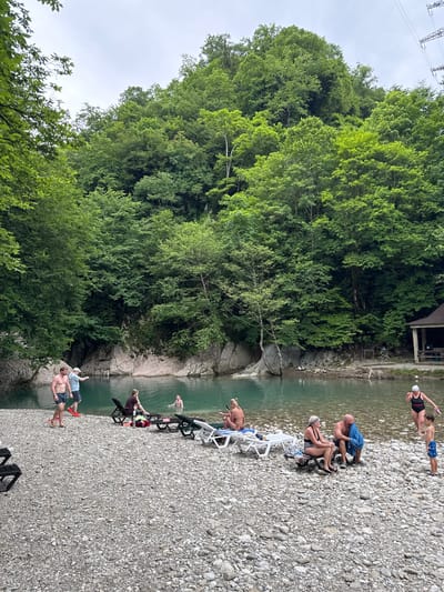 This image depicts a recreational area along a river in Khleborob, Russia. The scene is characterized by a clear, turquoise river flowing between rocky banks, with dense green forests covering the steep hillsides. On the pebble-strewn shore, several people are engaged in various activities.

In the foreground, a group of individuals are present. To the left, two men are standing, one pointing towards the water. Further down the shore, several people are seated on lounge chairs, relaxing. A few individuals are in the water, swimming or wading. One woman in a black swimsuit with a red strap is shown partially submerged in the water, with a young boy standing on the shore nearby.

On the right side of the image, a small, open-sided wooden structure with a brown roof is visible, possibly a changing room or shelter. The overall atmosphere suggests a tranquil summer day at a natural water resort.