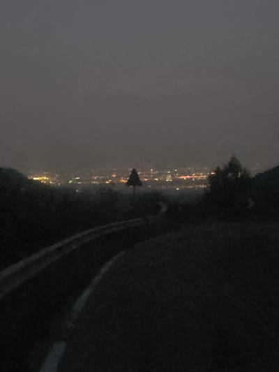The image depicts a dark, hazy view of a city at dusk or night, likely from an elevated vantage point. The lights of the city are visible in the distance, appearing as clusters of yellow and orange glows against the dark landscape. A road curves through the foreground, with white lines indicating lanes. A guardrail runs along the edge of the road. A triangular traffic sign is visible on the right side of the road in the middle ground. The sky is a uniform dark gray, suggesting overcast conditions or twilight. The overall impression is one of a quiet, somewhat isolated view of an urban area from a distance. The location is identified as Fumane, Italy.