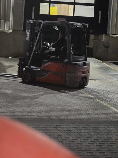 A forklift is parked on a paved area with a man seated in the driver's seat, appearing to be operating the vehicle. The forklift is red and grey, with visible forks. In the background, there is a building with windows and a dark exterior wall, suggesting an industrial or warehouse setting. The ground is paved with rectangular bricks. The scene is dimly lit, indicating it is nighttime or indoors with artificial lighting. The location is identified as Zeppelinheim, Germany.