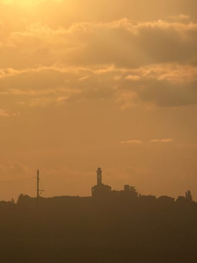 The image depicts an orange sky at sunset in Santo Stefano Belbo, Italy. Silhouetted against the sky are a line of trees, a utility pole, and what appears to be a tower. The sky is filled with long, streaky clouds.