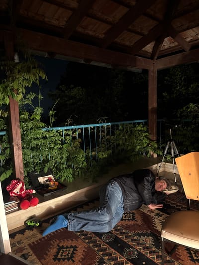 A man is lying on a rug in an outdoor setting in Kfar Vradim, Israel. He is wearing jeans, a black jacket, and blue socks. His eyes are closed. His hands are raised near his face. A chair is partially visible above him. A metal fence and plants are visible in the background. Toys are scattered near the edge of the rug. A wooden structure borders the scene on one side. It is nighttime.