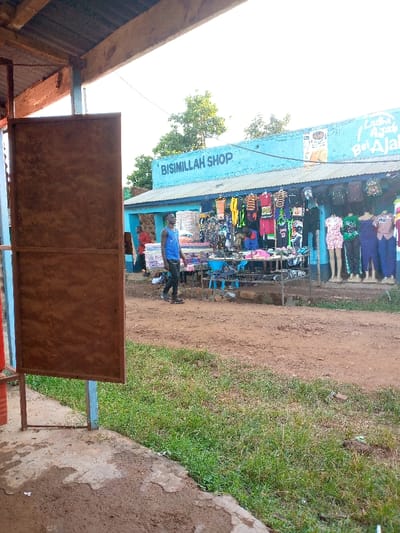A street scene shows a blue building with "Bismillah Shop" painted on the front. Clothing and mannequins are displayed under the awning. A man in a blue tank top walks past the shop. A rusty metal structure stands in the foreground on the left. The ground is a mix of dirt and green grass. The scene is bathed in daylight.