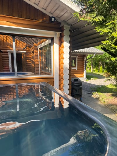 This media file shows an outdoor scene in Saue, Estonia, on a sunny day. The primary subjects are a rustic wooden cabin and a large, dark, reflective object that appears to be a hot tub or swimming pool. The cabin has a distinctive log construction with rounded, decorative trim along the roofline. A window on the cabin reflects the interior, which also seems to be made of wood.
In the background, a gravel path leads away from the cabin through a lush green garden with tall trees. Sunlight filters through the leaves, casting dappled shadows. A black portable speaker is positioned on the ground near the cabin.
The reflective surface of the hot tub or pool dominates the foreground and the right side of the image. It shows distorted reflections of the cabin, the trees, and the sky. Two dark silhouettes of birds are visible as reflections on the cabin window, possibly indicating decals or actual birds.
The overall impression is one of a tranquil, natural setting, likely a backyard or a vacation property, where one might relax outdoors. The bright sunlight suggests it is daytime. No visible text is present in the image.