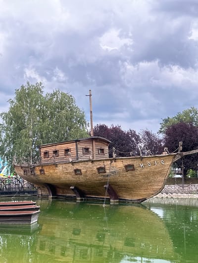 The image depicts a wooden ship structure situated in a pond with green water. The ship, which appears to be an attraction, has the word "HOJE" inscribed on its side. Several small boats are moored to the left of the ship. In the background, there are trees with lush green and dark red foliage, and a cloudy sky. A bridge is visible to the far left. The ship is positioned on land, with its hull resting on supports. This scene is located in the city of Dijelovi, Bosnia and Herzegovina, likely within a park or recreational area.