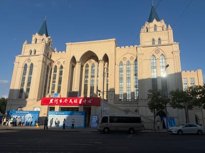 The image shows the exterior of a large, light-colored, castle-like building in 幸福乡, China. The building features multiple towers with pointed blue roofs, Gothic-style arched windows, and decorative stonework. In front of the building, there are several banners and signs, including a large red banner with Chinese characters. A group of people are gathered in front of blue informational displays, some appearing to be participants in an event. A silver minivan and a white sedan are parked on the street in front of the building. Trees line the sidewalk. The sky is clear and blue, and the sun is casting shadows.