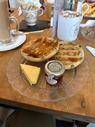 A close-up image depicts a meal served on a light-colored wooden table in a cafe or tea room located in Ambleside, United Kingdom. The central focus is a clear glass plate with an embossed floral edge, holding two toasted, currant-studded tea cakes or fruit buns. Adjacent to these are a triangular wedge of bright yellow butter and a miniature jar of Wilkin & Sons Tiptree Strawberry Preserve (28g). In the background, to the left, a tall glass mug contains a frothy, light brown beverage, resting on a white saucer with a spoon. Further back on the left, a decorative white teacup holds several brown and white sugar packets on a dark, irregularly shaped coaster. To the right, a white ceramic mug with a visible "Cottage Cafe Mr. H's" logo contains a dark beverage topped with cocoa powder. Additional blurry white crockery and food items are visible in the distant background, suggesting other patrons or table settings. The table itself displays a distinct wood grain and a visible seam. No individuals are directly visible in the frame, though the setting implies immediate human presence.