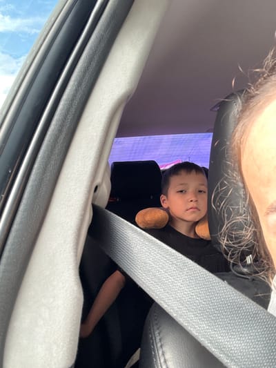 The image displays the interior of a vehicle, located in Sukhtelinskiy, Russia, during daytime. In the foreground, on the right, the partial face of an adult is visible, showing the right cheek, ear, and brown, curly hair. A grey seatbelt diagonally crosses the lower right, indicating the individual is seated in a front position.

Seated in the rear of the vehicle, slightly to the left of the center, is a young boy with dark hair, wearing a black t-shirt. He maintains a neutral expression and looks directly at the camera. Two brown, padded, spherical objects are positioned on either side of his head, suggesting a neck or head support. His left arm is visible, resting on a dark surface.

The car's interior features light grey and black trim on the left-hand side, corresponding to the door pillar. Through the upper left window, a clear blue sky with white clouds is visible. The rear window displays a distinct purple hue with horizontal lines, possibly a reflection or a digital screen. Dark-colored seating upholstery is present, along with a grey seatbelt secured around the child. The upper right portion of the image shows part of the vehicle's ceiling.