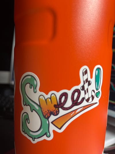 A sticker that reads "Sweet!" is adhered to the side of an orange thermos. The sticker features a green, dripping "S," the word "sweet" in a colorful, stylized font, a candy cane, and an exclamation point with a green outline. The thermos is on a dark surface, and the background is blurred. The image is taken from a close-up perspective. The specific location in Gresham, United States of America, cannot be determined from the provided image.