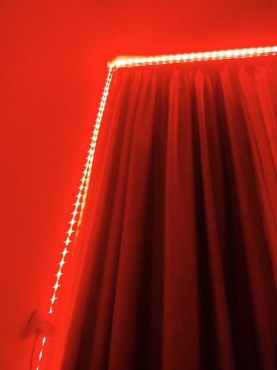 The image shows a close-up view of a red curtain with red LED strip lights. The lights are arranged along the top edge of the curtain and a vertical strand runs down the left side, illuminating a wall and a white electrical outlet. The scene is set in a room with red walls. There are no people or animals visible in the image. The lighting is entirely red, creating a warm and dramatic atmosphere. The specific location in the city of Criuleni, Moldova cannot be determined from the visual information provided.
