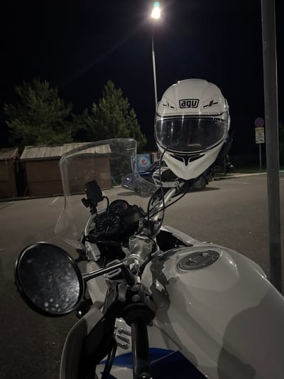 A white AGV motorcycle helmet sits atop a white motorcycle in a dimly lit outdoor area, likely a parking lot, at night. The motorcycle's handlebars, mirrors, and instrument cluster are visible. In the background, there are trees, a wooden structure resembling a shed or kiosk, and a street lamp emitting light. A street sign is also visible on the right side of the image. The scene appears to be in the city of Privolzhsky, Russia, based on the prompt. There are no people visible in the image.