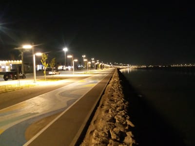 The image depicts a nighttime scene along the waterfront in Al Khobar, Saudi Arabia. A paved walkway with decorative colored patterns runs alongside a road. Streetlights illuminate the area, casting warm light on the path and the adjacent water. The sea is visible on the right side of the image, reflecting the distant lights of the city. On the left, cars are seen driving on the road, and buildings with illuminated facades are present in the background. A rocky embankment separates the walkway from the water. The overall impression is of a well-lit, modern urban waterfront at night.