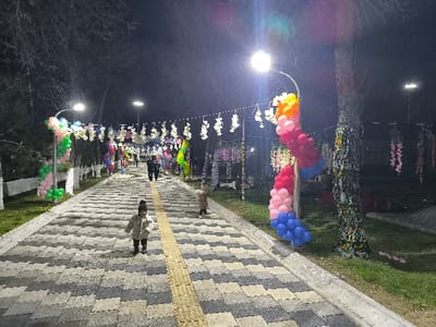 The image displays a nighttime scene within a decorated public pathway in Pakhtakor, Uzbekistan. A paved pedestrian path, constructed from grey and black interlocking pavers and featuring a central yellow tactile strip, extends into the background. The area is illuminated by multiple bright street lamps. In the immediate foreground, a small child, likely a toddler, is standing centrally on the path, wearing a light-colored winter coat, dark pants, dark boots, and a dark knit hat. Another small child in a light-colored coat is partially visible on the right side of the path, near a tree. Further down the pathway, two adults in dark clothing walk away from the viewer, one appearing to accompany another small child.
The environment is adorned with numerous festive decorations. On the left, a white post is wrapped with a green and pink balloon garland. On the right, a street lamp post is decorated with a multi-colored balloon garland in shades of orange, yellow, pink, red, and blue. Overhead, strings of white floral or light decorations are suspended across the path. Bare trees line both sides of the pathway, and one tree trunk on the right is wrapped in colorful patterned fabric. Additional colorful fabric or artificial floral chains hang from tree branches and overhead lines. Grass strips border the paved pathway on both sides.