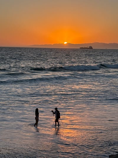 In Manhattan Beach, California, at dusk, two people are in the shallow ocean water near the shore. A person on the left, silhouetted against the orange sky, stands facing right, appearing to be looking at the person on the right. The person on the right, also silhouetted, is facing the person on the left and is holding a camera on a tripod. The sun is low on the horizon, casting a warm glow on the water and sky. A cargo ship is visible in the distance on the ocean. The waves are gentle, and the water reflects the colors of the sunset. The beach sand is submerged in the water.