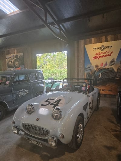 The image depicts an indoor vehicle display area in Ban Sahakon 2, Thailand. The environment features a dark, industrial-style ceiling with exposed metal trusses and a concrete floor. Walls are constructed from vertical wooden planks.

In the foreground, a light blue vintage convertible sports car is centrally positioned, facing slightly towards the left. It features round headlights, a chrome grille, chrome bumpers, multi-spoke silver wheels, and a black roll bar behind the cockpit. A white circular decal with the black number "54" is prominently displayed on its hood, with "54" also present on the side panel. A Thai license plate, partially legible as "DH 7165," is affixed to the front bumper.

To the left of the sports car, a dark-colored (likely gray or black) vintage pickup truck or panel van is partially visible. Text on its side reads "SKYGGA."

In the background, an open doorway reveals an exterior scene dominated by lush green trees and foliage under daylight. Another vehicle, possibly light green, is partially visible beyond the doorway in the exterior setting.

Wall decor includes a large vertical poster on the right wall promoting the "Goodwood Festival of Speed," depicting vintage race cars and indicating a date of "23 June 1996." A smaller poster on the left wall displays a silver vintage racing car with the number "3." No individuals are present in the scene.