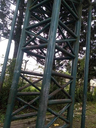 This image depicts a segment of a large, dark teal-painted tubular steel lattice structure situated in Namthoung, Laos. The structure dominates the foreground, viewed from a low angle looking upwards. It consists of prominent vertical and diagonal members forming a triangular truss pattern. The dark teal paint exhibits significant wear, fading, and chipping, revealing areas of underlying rust, particularly on the diagonal cross-members. Faint white markings, possibly alphanumeric, are visible on some vertical components.
Beyond the lattice structure, dense green vegetation is present on both sides. To the left, a mature coniferous tree with a thick, dark trunk stands against a bright, overcast sky. Below the tree, a section of a wire fence, potentially barbed, is visible amidst overgrown foliage. To the right, further trees and shrubs frame a partial view of a light-colored building or roof structure in the background. The ground beneath the lattice structure appears to be a mix of dirt and possibly concrete, with sparse green growth and a small piece of yellow debris. No individuals, actions, or direct interactions are visible within the frame. The ambient lighting suggests an overcast sky conditions.