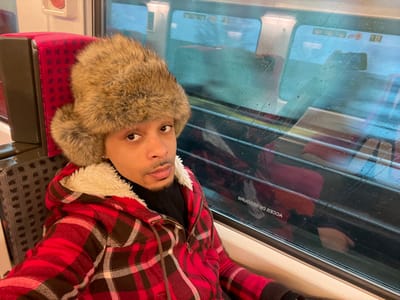 An adult male is seated inside a train carriage, located in the city of Saint-Cyr-l'École, France. He is positioned in the left and central foreground, looking directly at the camera. The individual has light brown skin, dark eyes, and a short beard and mustache. He is wearing a brown fur-lined ushanka hat, a dark-colored top, and a red and black plaid flannel jacket with a cream-colored fleece lining visible at the collar. To the left and behind the subject, a red patterned fabric train seatback is visible. To the right, a train window displays reflections of the carriage interior, including additional red seats and the inverted text "ACCES SECOURS." The view through the window is blurred and features blue tones, with water droplets visible on the glass exterior, suggesting the train is in motion or has been exposed to precipitation.