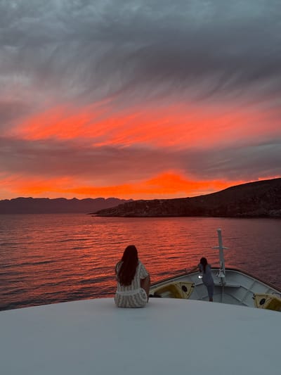 The image depicts a sunset view from a boat off the coast of Unknown, Mexico. The sky is filled with vibrant orange and gray clouds, casting a reddish glow on the water. A rocky coastline is visible in the distance. Two individuals are present on the boat. One is standing near a mast-like structure, while the other, with long hair, is closer to the edge of the boat. The boat is white and has a lifeboat. The environment suggests a calm sea and a dramatic sunset.