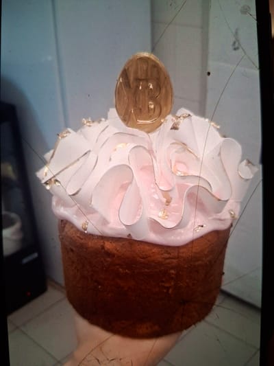 The media file shows a close-up, angled shot of a decorated pastry, likely a cake or a large cupcake, placed on a light-colored surface. The pastry has a rich brown base, likely cake, topped with a generous swirl of pale pink frosting. The frosting is decorated with delicate gold leaf accents, giving it a luxurious appearance. Attached to the side of the pastry is a circular, gold-colored object with an embossed design, possibly a coin or a decorative topper, featuring a stylized face or symbol. The background is out of focus but appears to be an indoor setting with neutral-colored walls or surfaces. The image has a slightly grainy texture and visible scratches and dust on the surface it was captured on, suggesting it might be a photograph of a screen or a poorly maintained camera lens. There is no visible text in the image. The lighting appears to be artificial and diffused. The overall impression is that of a celebratory or decorative pastry, possibly prepared for a special occasion. The location context of Medvezhyi Ozyora, Russia, is not directly evident from the visual information.