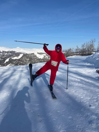 An individual is depicted performing an athletic pose on a snow-covered ski slope, situated in Mušovića Rijeka, Montenegro. The subject wears a bright red, hooded, full-body jumpsuit with a light-colored rectangular patch on the upper torso. Essential gear includes a black ski helmet and purple-tinted ski goggles, through which a smile is visible. The person holds two black ski poles; the left pole is raised high with the arm bent, while the right pole is held lower near the hip. The individual is balanced on their right ski, with the left ski lifted vertically off the snow, supported by a bent left leg. The immediate environment is a groomed ski trail exhibiting ski tracks and strong shadows, including a prominent shadow of the person taking the photograph. The background features extensive snow-covered mountainous terrain with sparse tree cover under a clear blue sky displaying faint contrails.