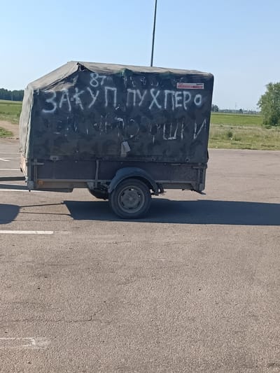 A dark-colored trailer with a covered top is parked on an asphalt surface. The cover has graffiti in white paint that appears to be in the Russian language. In the background, there is a grassy field and some trees. The location appears to be a rural or semi-rural area with a clear sky. The trailer is in Алтынды ауылдық округі, Kazakhstan.