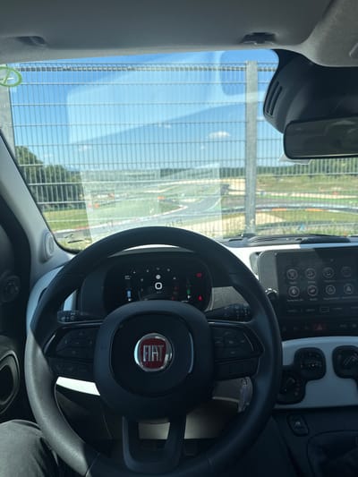Here is a description of the image:

The image shows the interior of a Fiat car, specifically the steering wheel and dashboard. The steering wheel has the Fiat logo in the center and various control buttons. The dashboard includes a digital display with vehicle information and a multimedia system screen. Through the windshield, a view of an outdoor area is visible, possibly a racetrack, based on the structures and layout seen in the background.

The image shows Campagnano di Roma, Italy.