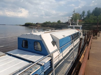 The image depicts a side view of a white and blue passenger boat docked at a pier. The boat is moored on a wide, calm body of water, with a shoreline visible in the background lined with trees and debris. The sky is partly cloudy. The pier is made of concrete and metal structures, with some visible rust. The boat appears to be older and in a state of disrepair in some areas, with weathered paint and visible wooden planks. The markings "PTO 50 58" are visible on the boat's superstructure. The overall impression is one of a quiet, possibly industrial, waterfront setting. There are no people visible in the image. The location is identified as Alexandrovskoe, Russia.