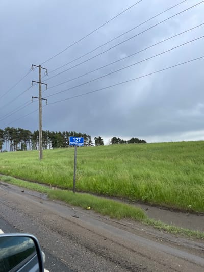 The image depicts a roadside scene near Kokino, Russia. A sign marked "127 РОСАВТОДОР" is visible beside a narrow paved road. Adjacent to the road is a field of green grass. A row of evergreen trees lines the horizon. In the background is a cloudy, overcast sky. A utility pole with power lines is on the left side of the road. The image appears to have been taken from inside a vehicle, with a side mirror partially visible in the bottom right corner.