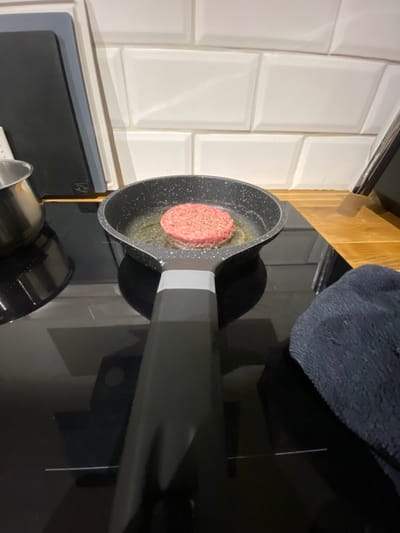 A raw hamburger patty sizzles in a black speckled pan on a glass stovetop. Cooking oil bubbles around the edges of the patty. The pan's handle extends horizontally towards the right. A dark gray cloth rests near the back of the stovetop. White subway tiles line the wall adjacent to the stovetop. A stainless steel object sits below the pan.