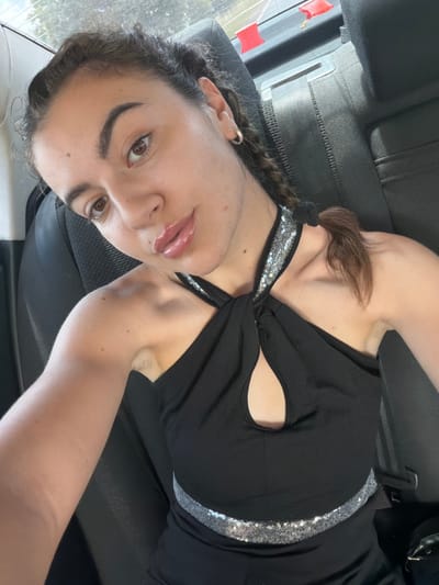A casual selfie features a young woman reclined in what appears to be the back seat of a vehicle, likely a car or bus, in Sadievo, Bulgaria. She is positioned with her head tilted, looking directly into the camera. Her dark hair is styled with a braid and secured with a black hair tie, with some strands visible around her face. She wears a black top or dress with a distinct design, featuring a twisted neckline, a cut-out detail on the chest, and a silver sequined strap or trim running across the garment. Her lips are enhanced with a glossy product, and her eyebrows are well-defined.

The background consists of dark, possibly leather or fabric, vehicle upholstery and parts of the car interior. A seatbelt buckle is clearly visible to her left. Through a window in the upper left of the frame, there's a glimpse of an outdoor scene, with two small red flag-like objects visible, suggesting a journey or stop. Natural light illuminates the scene, casting some shadows and indicating it is daytime. The woman has a calm and direct expression. No visible text is present in the image.