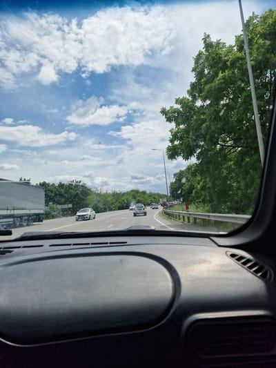 The image is taken from inside a vehicle, looking forward through the windshield. The scene is a highway with several cars and a large truck visible on the road. The sky is blue with scattered white clouds, and lush green trees line the side of the highway. The dashboard of the vehicle is partially visible in the foreground. The overall impression is of a journey on a highway on a cloudy day. The location is described as Zmeyka, Russia, though there are no specific visual cues in the image to confirm this.