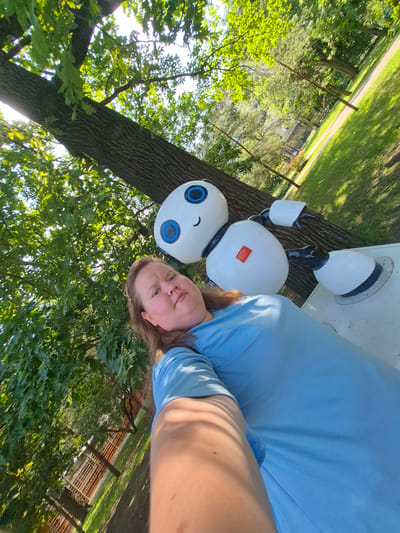In a park setting in Pavlovsk-2, Russia, a woman in a light blue t-shirt is taking a selfie. She is positioned in the foreground, with her arm outstretched holding the camera. To her right, and slightly behind her, is a white, cartoonish robot statue with a black neck, black arms, and large blue and black eyes. The robot has a small red square on its chest. The background consists of a large tree trunk to the left, and lush green foliage, grass, and a pathway in the distance. Sunlight filters through the leaves, creating dappled light. A wooden fence is visible to the lower left of the frame.