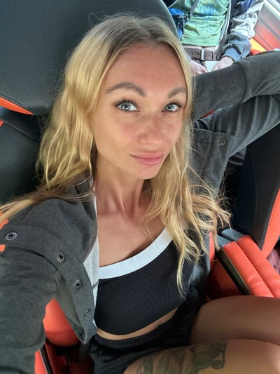 A woman with blonde hair and blue eyes is photographed from the chest up. She is wearing a dark gray hooded jacket over a black ribbed crop top with a white trim, and black shorts. Her left arm is bent and resting behind her head, with her hand near the back of her head. She is seated in what appears to be a car or a racing-style chair, with black upholstery and orange accents. The background shows parts of another person seated behind her, wearing a green shirt and a brown belt. The image appears to be a selfie, taken indoors or in a vehicle, with no discernible external environmental elements related to Phan Son, Vietnam.