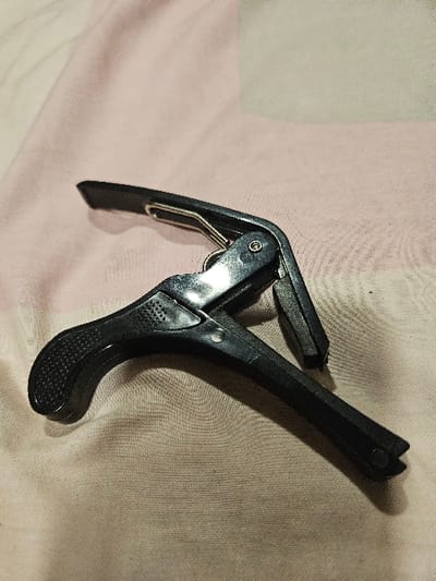 A black guitar capo is positioned diagonally from the lower-left to the upper-right on a light pinkish-beige, finely textured fabric surface. The capo is constructed primarily of black plastic with visible metallic components, including a silver-toned spring mechanism and hinge pins. The lower handle features a textured, stippled grip pattern. The jaws of the capo are slightly open, revealing a dark, soft padding on the inner surfaces. The lighting casts subtle shadows and highlights on the object and fabric. This scene is located in the city of Padajaya, Indonesia.